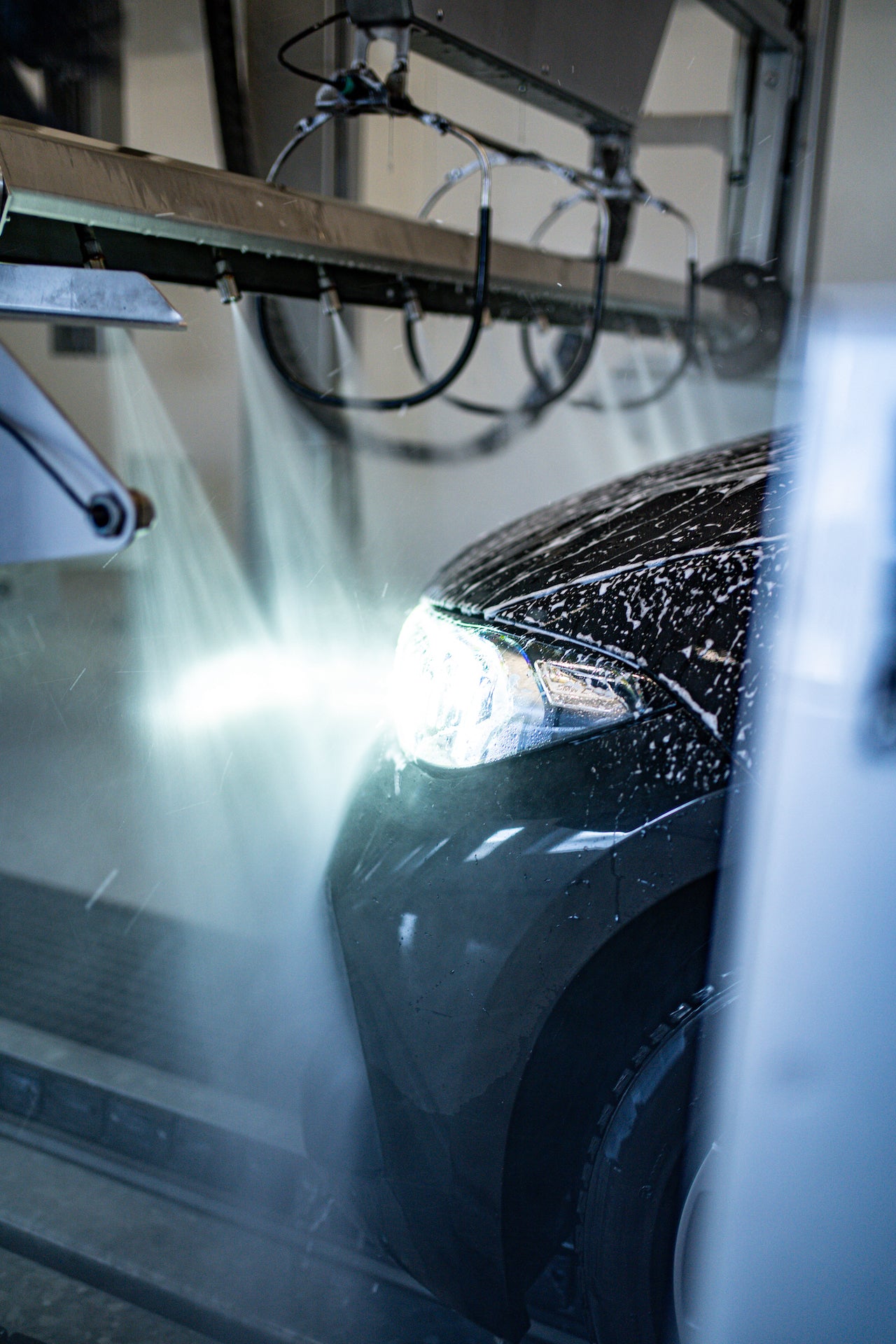 Eine Fahrzeugfront von einem dunklen Auto ist mit Schaum benetzt und wird mit Hochdruckdüsen abgespült
