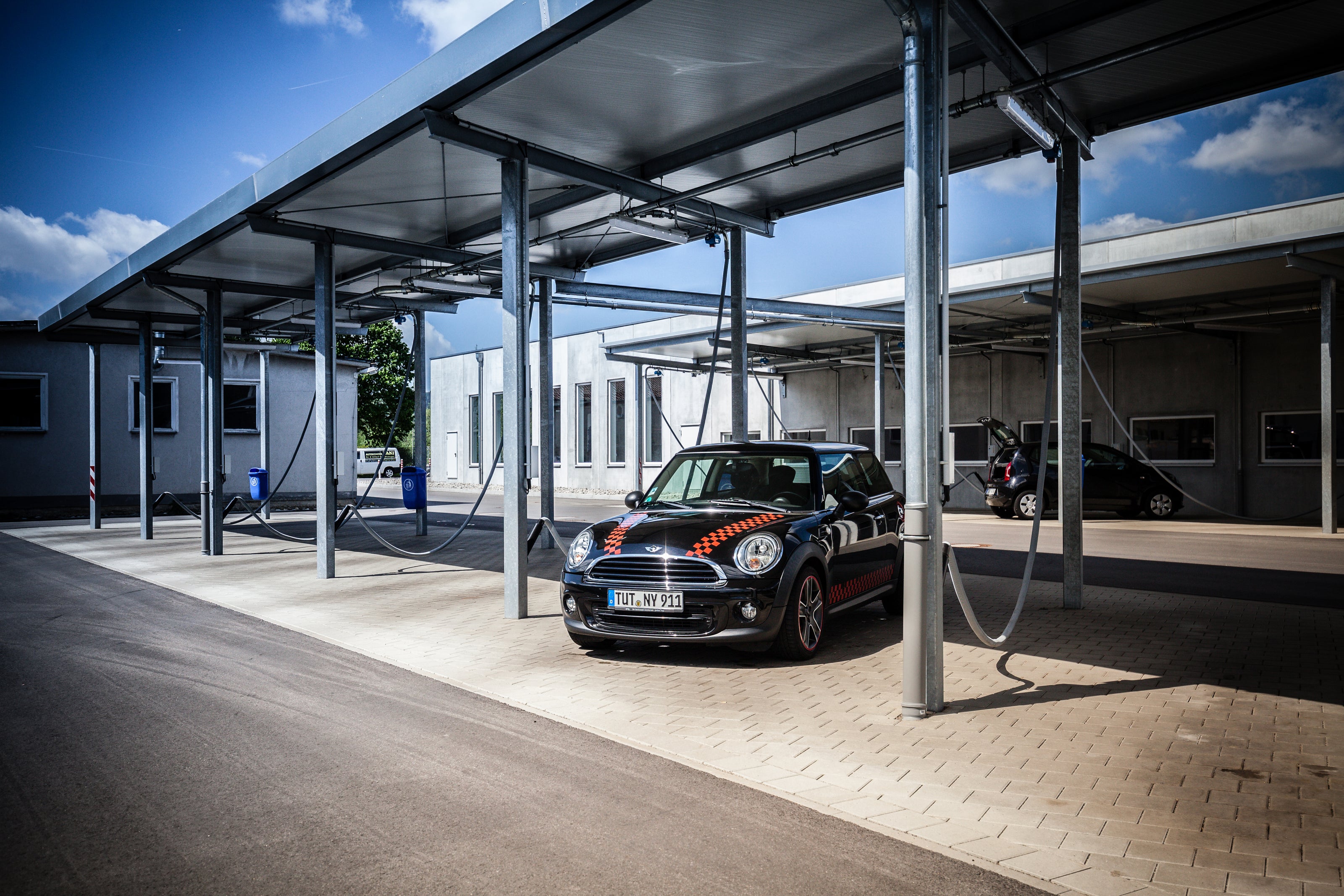 Schwarzer Mini Cooper mit orange-weißem Muster an der Motorhaube steht auf den Saugerplätzen an der Waschstraße unter einer Überdachung auf gepflastertem Platz.