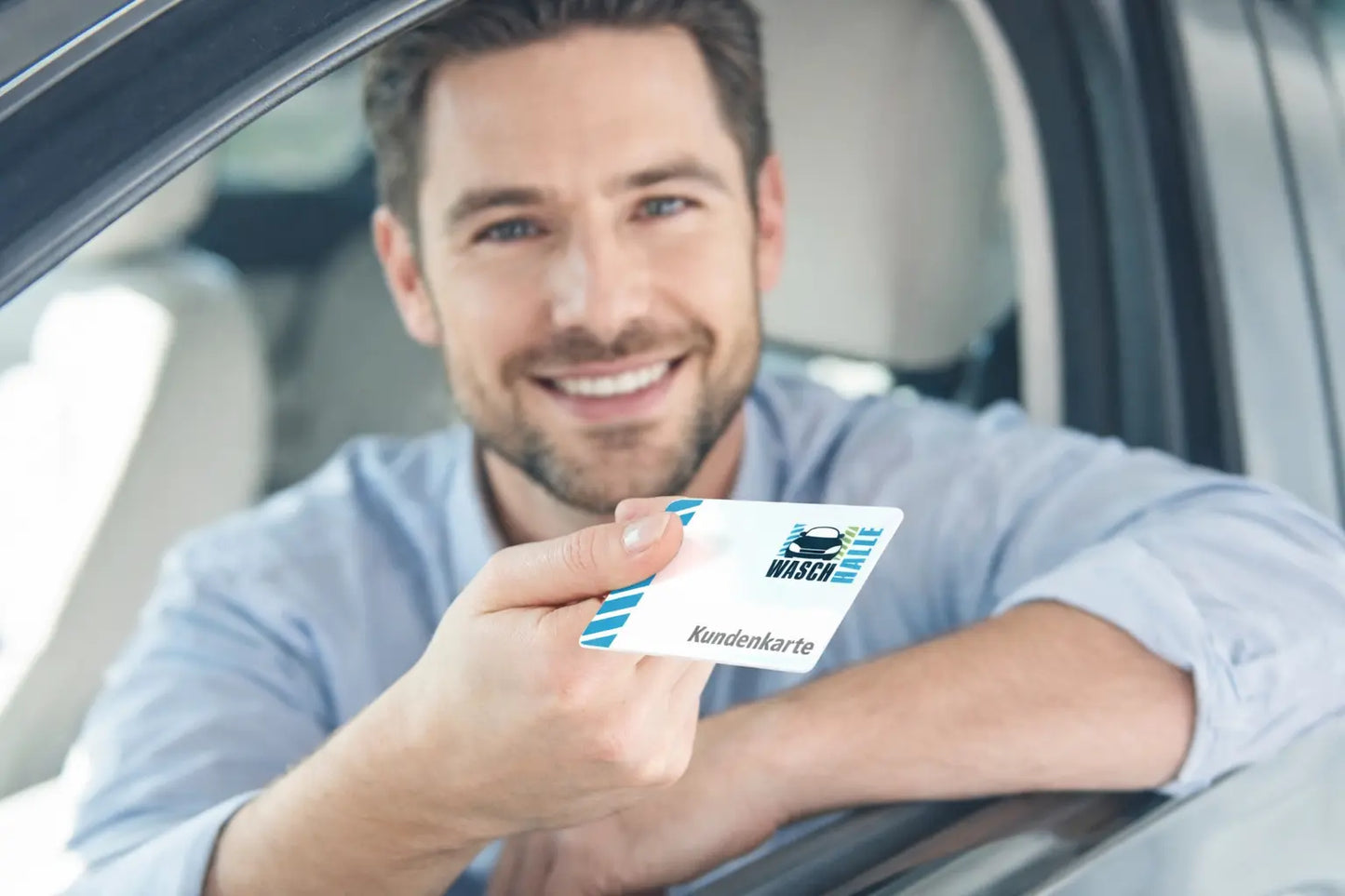 Ein lächelnder Mann sitzt in einem Auto mit heruntergelassenem Fenster und hält eine weiße „WASCHHALLE“-Kundenkarte hoch.