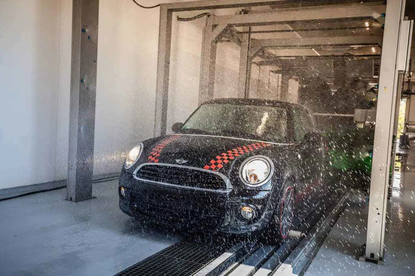 Ein schwarzer Mini Cooper mit rot karierten Streifen fährt durch eine automatische Waschanlage, umgeben von spritzendem Wasser und Maschinen in einer Autowaschanlage.