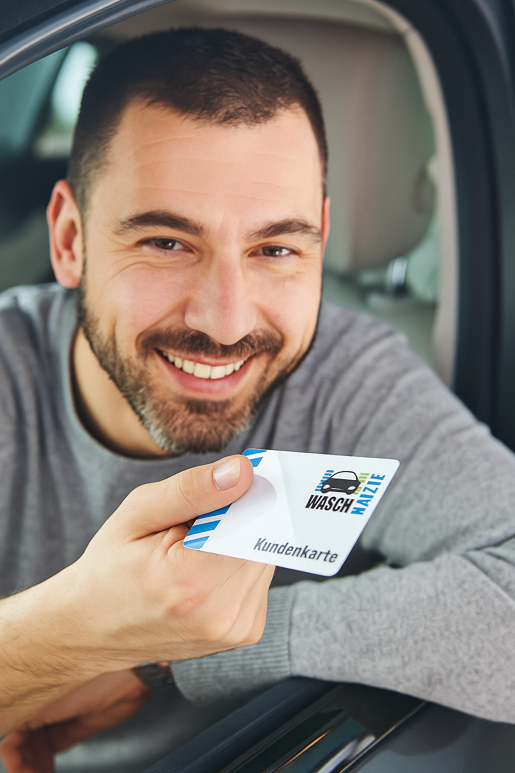 Ein lächelnder Mann sitzt in einem Auto mit heruntergelassenem Fenster und hält eine weiße „WASCHHALLE“-Kundenkarte hoch.