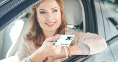 Ein lächelnde Frau sitzt in einem Auto mit heruntergelassenem Fenster und hält eine weiße „WASCHHALLE“-Kundenkarte heraus.
