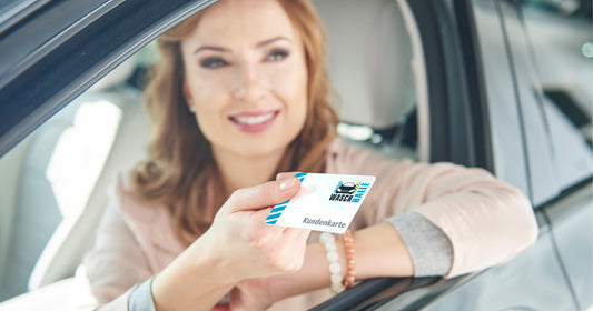 Ein lächelnde Frau sitzt in einem Auto mit heruntergelassenem Fenster und hält eine weiße „WASCHHALLE“-Kundenkarte heraus.