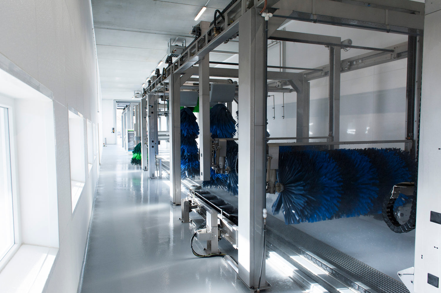 Eine moderne, leere automatische Autowaschanlage mit großen rotierenden blauen und grünen Bürsten, Metallrahmen und heller Beleuchtung, die sich auf dem sauberen, glänzenden Boden spiegelt.