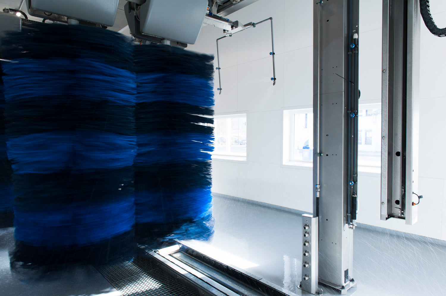 Große blaue und schwarze rotierende Bürsten in einer automatischen Waschstraße, mit spritzendem Wasser und weißen Kachelwänden im Hintergrund. Helles natürliches Licht fällt durch die Fenster.