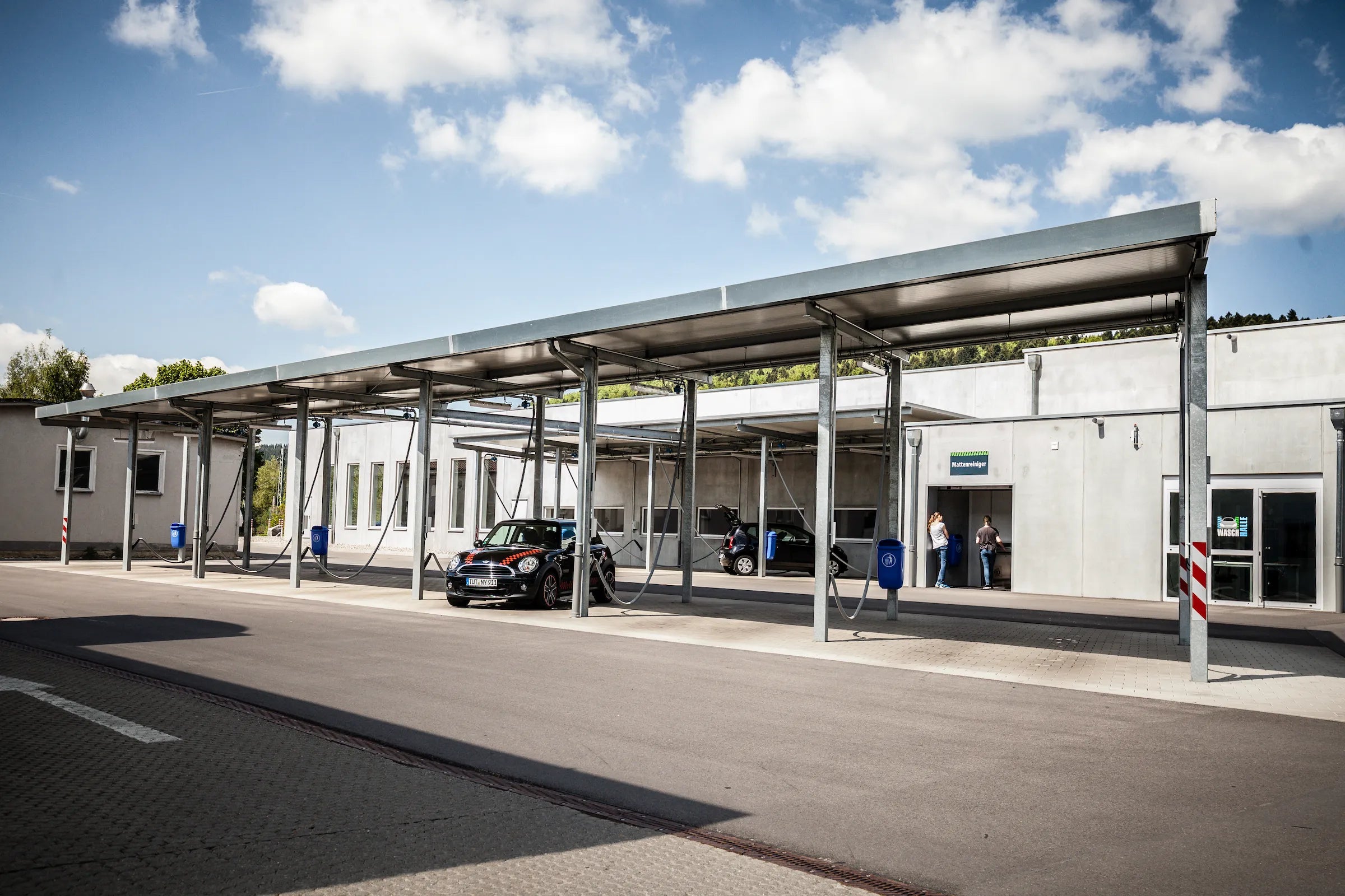 Ein modernes Gebäude mit einer Carport-Struktur aus Metall bietet an einem sonnigen Tag Schutz für geparkte Autos. Zwei Personen stehen in der Nähe des Eingangs, und der Himmel ist teilweise bewölkt. Die Umgebung scheint sauber und ordentlich zu sein.