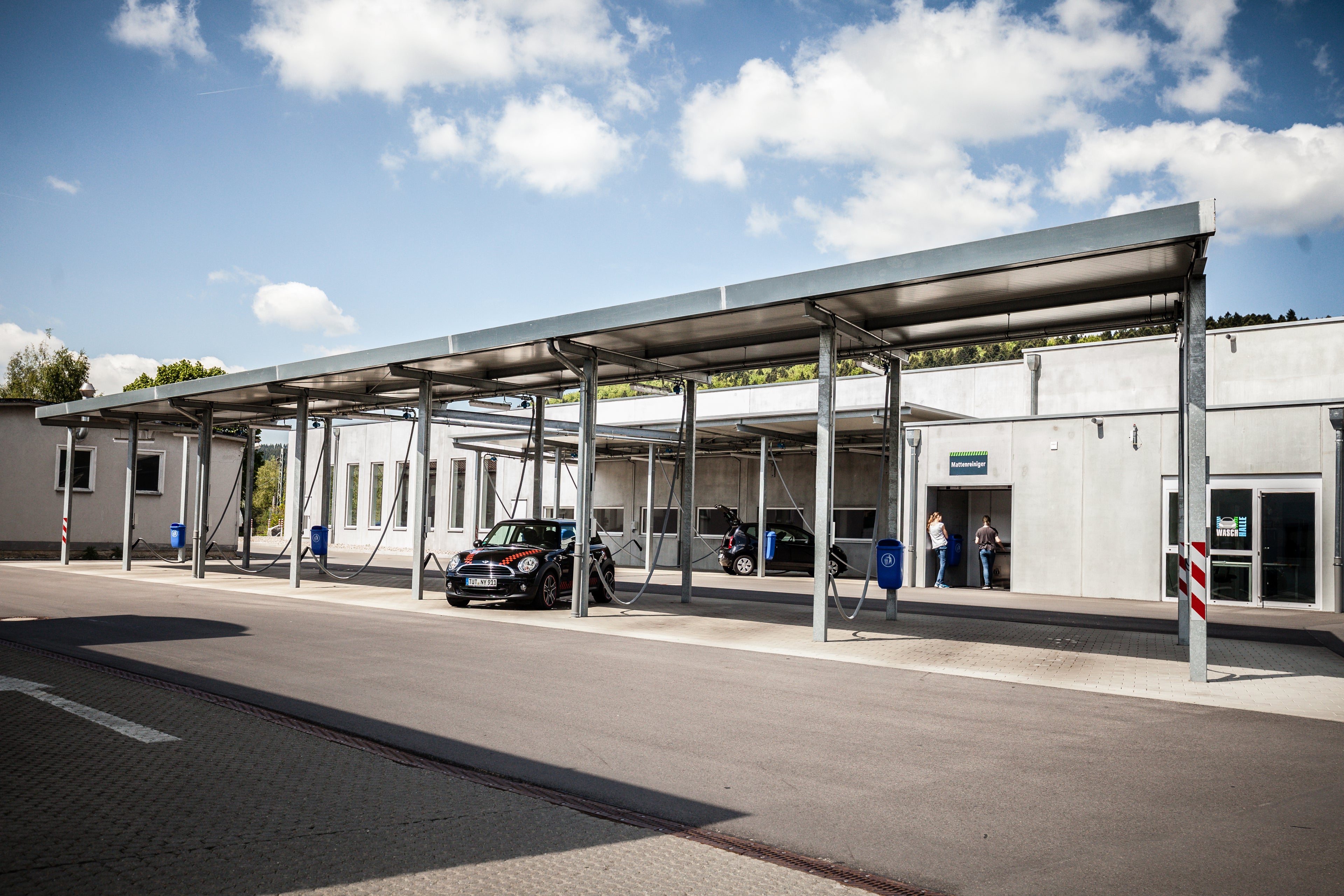 Ein modernes Gebäude mit einer Carport-Struktur aus Metall bietet an einem sonnigen Tag Schutz für geparkte Autos. Zwei Personen stehen in der Nähe des Eingangs, und der Himmel ist teilweise bewölkt. Die Umgebung scheint sauber und ordentlich zu sein.