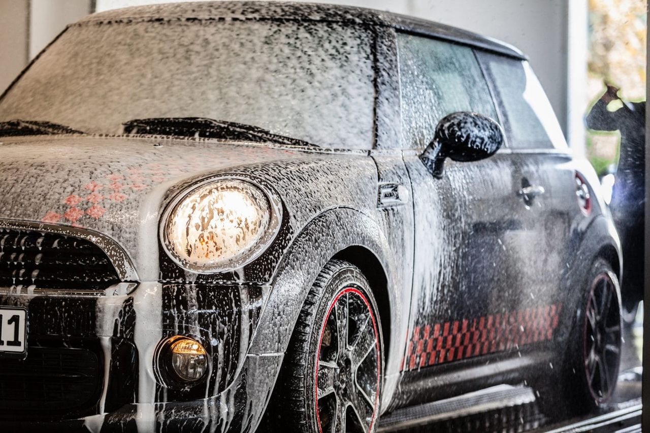 Schwarzer Mini Cooper mit roten Karomustern, bedeckt mit weißem Schaum beim Autowaschen.