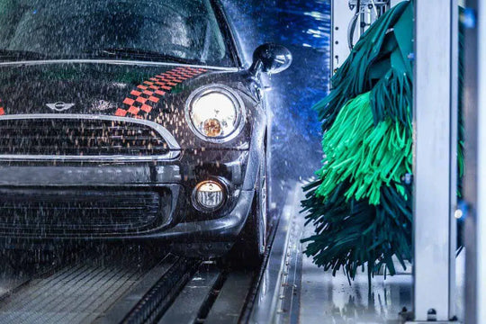 Ein schwarzer Mini Cooper mit rot karierten Streifen fährt durch eine automatische Waschanlage, in der Wasser versprüht und grüne Bürsten die Fahrzeugseite reinigen. Die Scheinwerfer des Autos sind eingeschaltet.