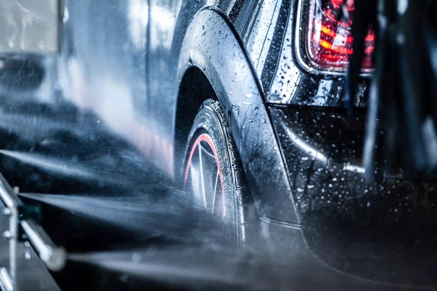 Nahaufnahme eines schwarzen Autos, das in einer automatischen Waschanlage gewaschen wird, wobei die Wasserdüsen die Räder und den hinteren Teil besprühen. Das Auto ist nass und glänzt, mit Wassertropfen auf der Oberfläche.